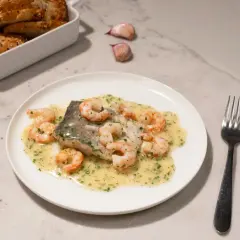 Deliciosa merluza con gambas en salsa cremosa, acompañada de pan rústico y ajos.
