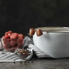 Preparación para un delicioso fondue de carne: trozos de carne cruda, ajo, granos de pimienta y una olla con caldo humeante.