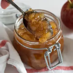 Cuchara sirviendo mermelada de manzana casera de un frasco. Manzanas rojas frescas al lado.