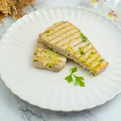 Dos filetes de atún a la plancha, con marcas de parrilla y aderezados con ajo y perejil, servidos en un plato blanco con un tenedor.