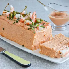 Pastel de pescado decorado con gambas, pepinillos y alcaparras. Servido en una bandeja blanca con una porción cortada.