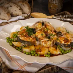 Merluza a la gallega arguiñano, jugosos trozos de pescado con patatas y guisantes, servidos en una fuente decorada.