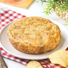 Tortilla con patatas de bolsa, dorada y jugosa, servida en plato blanco sobre mantel de cuadros rojos. Patatas fritas crujientes alrededor.