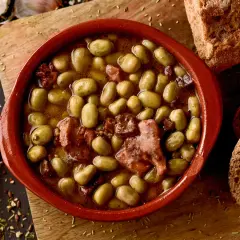 Un cuenco de barro con habas a la catalana, trozos de tocino y pan rústico al lado.