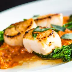 Deliciosas vieiras a la plancha, doradas y jugosas, servidas sobre una cremosa salsa naranja con brócoli tierno y rodajas de chorizo.