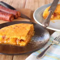 Deliciosa tortilla de chorizo, jugosa y dorada, servida en plato oscuro. Chorizos crudos al fondo.