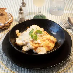 Delicioso bacalao a la nata gratinado, servido en un cuenco negro, acompañado de pan rústico y cubiertos.