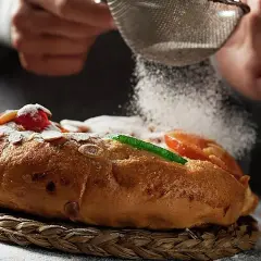 Manos espolvorean azúcar glas sobre un roscón de reyes decorado con frutas escarchadas y almendras. ¡Una delicia para tu roscón de reyes receta!