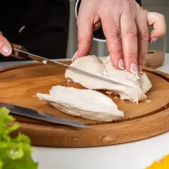 Manos cortando pechuga de pollo hervido en tabla de madera. El tiempo de cocción del pollo hervido es clave para su jugosidad.