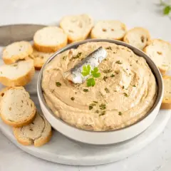 Un bol de pate de sardinas cremoso, decorado con una sardina y perejil, rodeado de rebanadas de pan tostado.