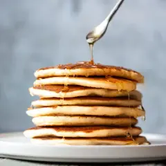 Una pila de tortitas sin mantequilla, doradas y esponjosas, con sirope cayendo de una cuchara.
