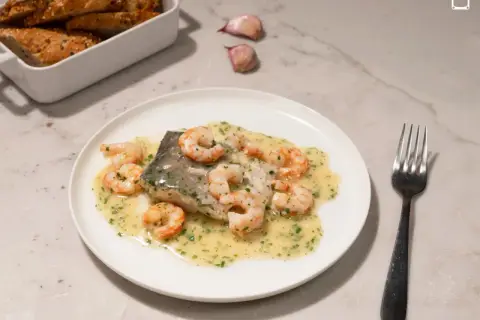 Deliciosa merluza con gambas en salsa cremosa, acompañada de pan rústico y ajos.
