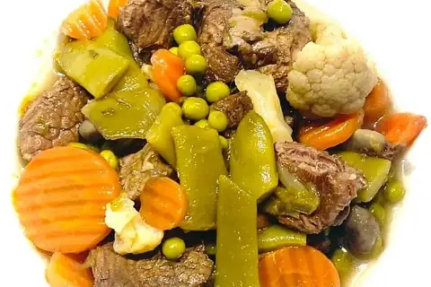 Ternera a la jardinera con trozos de carne tierna, zanahorias, guisantes, pimientos verdes y coliflor en un plato blanco.