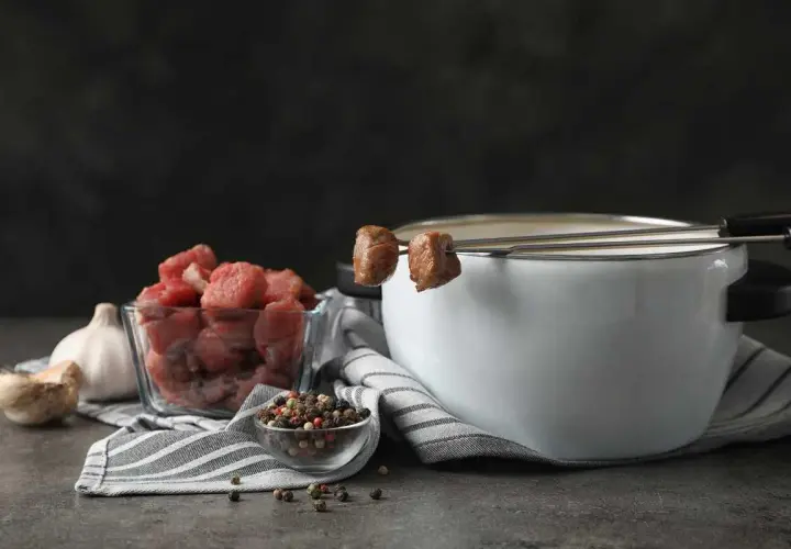 Preparación para un delicioso fondue de carne: trozos de carne cruda, ajo, granos de pimienta y una olla con caldo humeante.