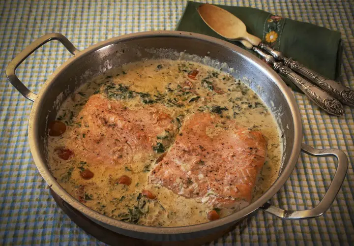 Dos filetes de salmón a la toscana en una sartén con salsa cremosa, espinacas y tomates cherry.