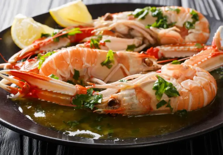 Cigalas a la plancha, jugosas y doradas, servidas con perejil picado y gajos de limón en un plato oscuro.