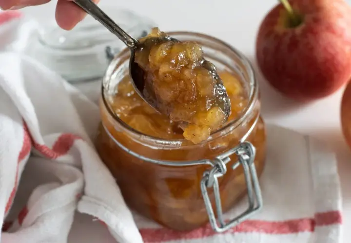 Cuchara sirviendo mermelada de manzana casera de un frasco. Manzanas rojas frescas al lado.