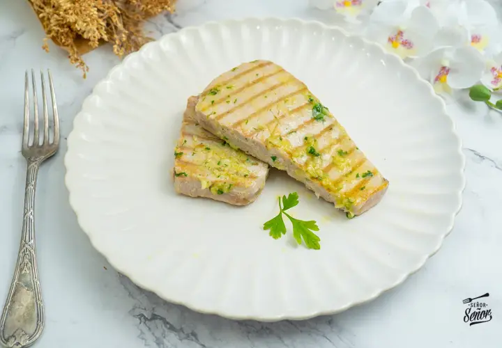 Dos filetes de atún a la plancha, con marcas de parrilla y aderezados con ajo y perejil, servidos en un plato blanco con un tenedor.