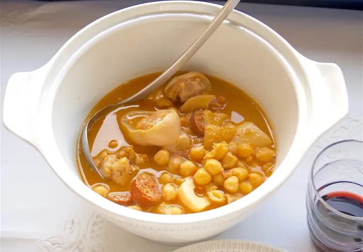 Un plato humeante de callos a la gallega, con garbanzos, chorizo y trozos de carne en una cazuela blanca.