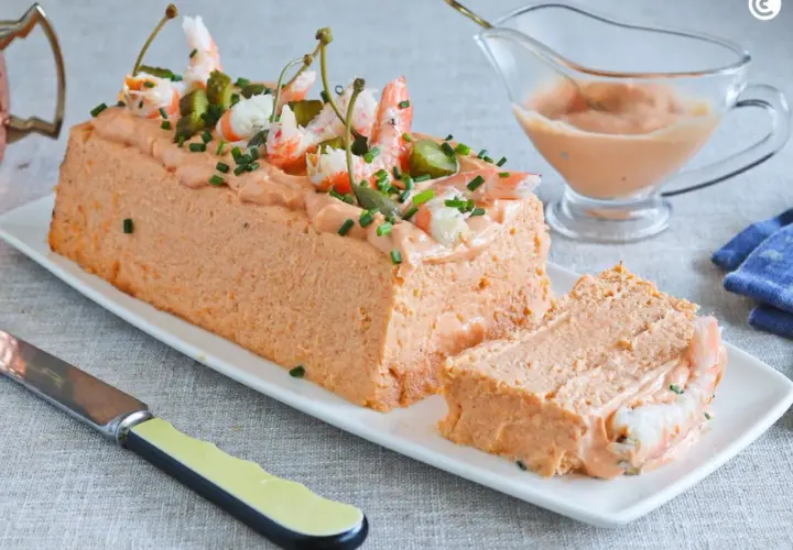 Pastel de pescado decorado con gambas, pepinillos y alcaparras. Servido en una bandeja blanca con una porción cortada.