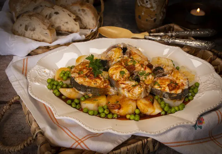 Merluza a la gallega arguiñano, jugosos trozos de pescado con patatas y guisantes, servidos en una fuente decorada.