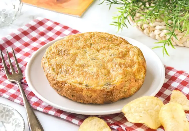 Tortilla con patatas de bolsa, dorada y jugosa, servida en plato blanco sobre mantel de cuadros rojos. Patatas fritas crujientes alrededor.