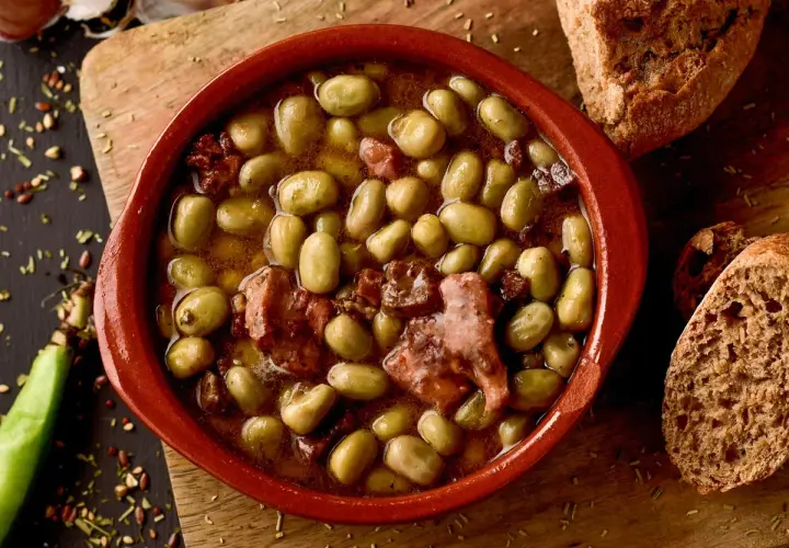 Un cuenco de barro con habas a la catalana, trozos de tocino y pan rústico al lado.