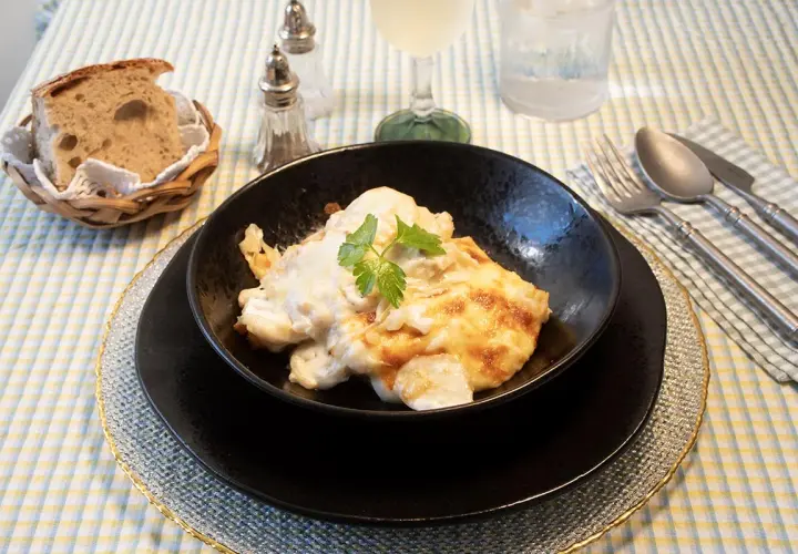 Delicioso bacalao a la nata gratinado, servido en un cuenco negro, acompañado de pan rústico y cubiertos.