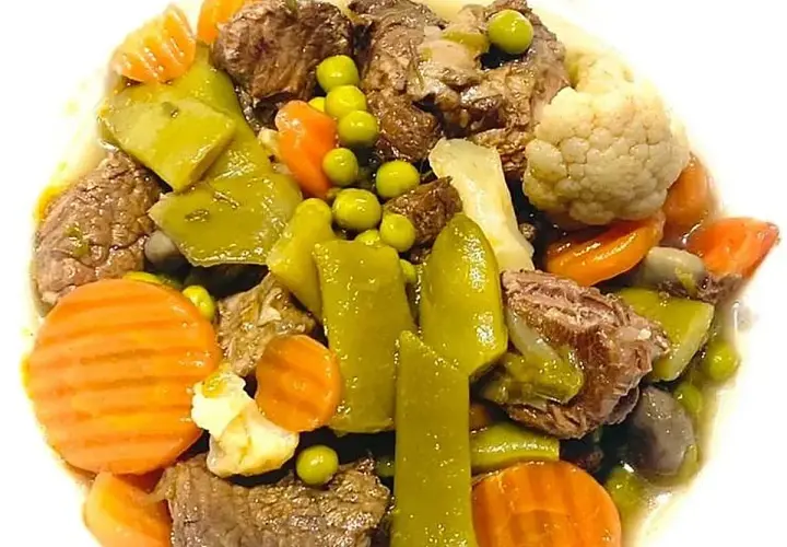 Ternera a la jardinera con trozos de carne tierna, zanahorias, guisantes, pimientos verdes y coliflor en un plato blanco.