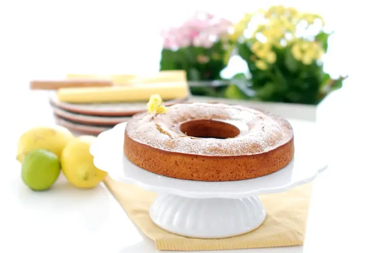 Bizcocho de limón Thermomix esponjoso y dorado, espolvoreado con azúcar glas. Limones y lima frescos al lado.