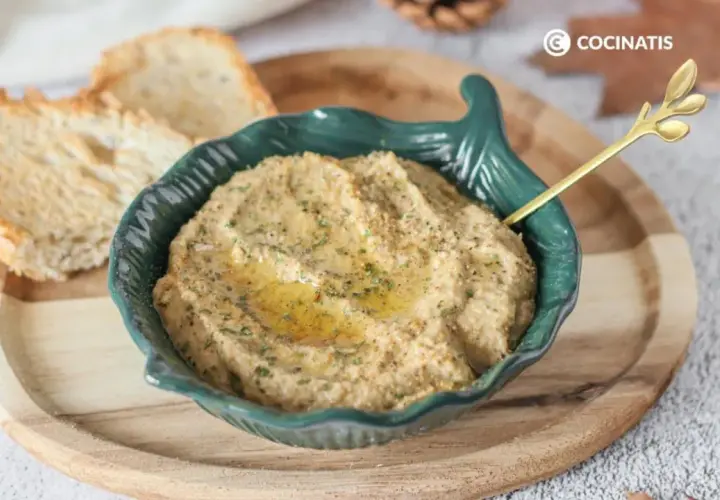 Delicioso pate de berenjena cremoso, servido en un cuenco verde con un toque de aceite de oliva y perejil. Acompañado de tostadas crujientes.