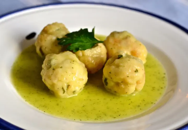 Cinco albóndigas de merluza caseras flotan en una salsa verde cremosa, adornadas con una ramita de perejil fresco.