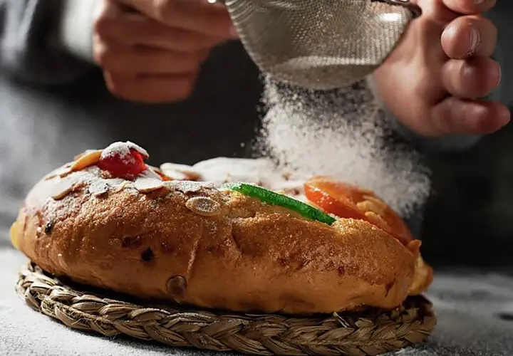 Manos espolvorean azúcar glas sobre un roscón de reyes decorado con frutas escarchadas y almendras. ¡Una delicia para tu roscón de reyes receta!