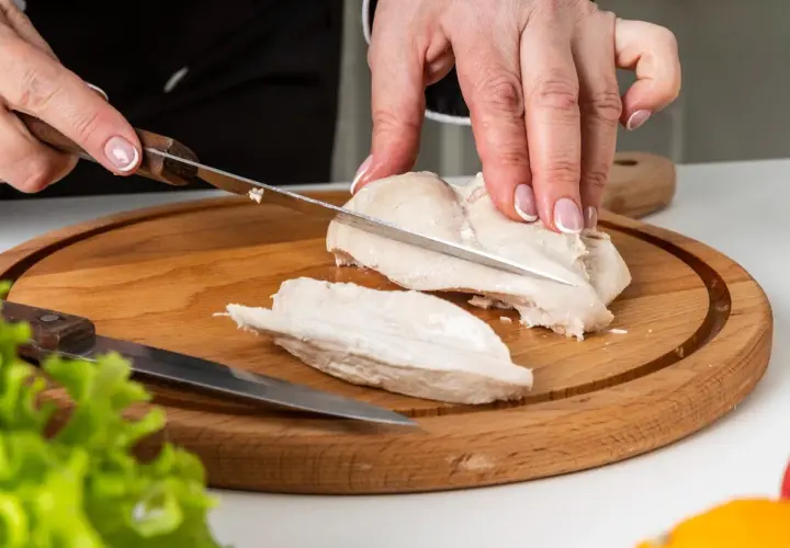 Manos cortando pechuga de pollo hervido en tabla de madera. El tiempo de cocción del pollo hervido es clave para su jugosidad.