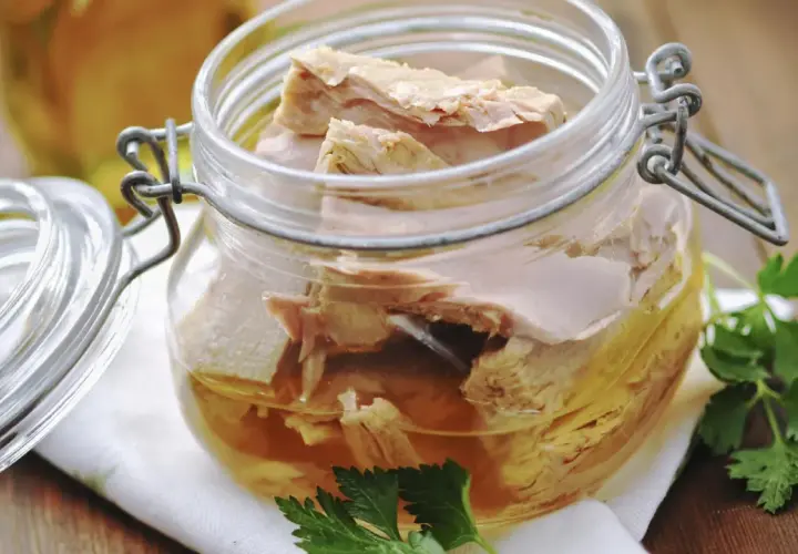 Un frasco de vidrio con atún en escabeche, listo para disfrutar. Unas hojas de perejil adornan la escena.