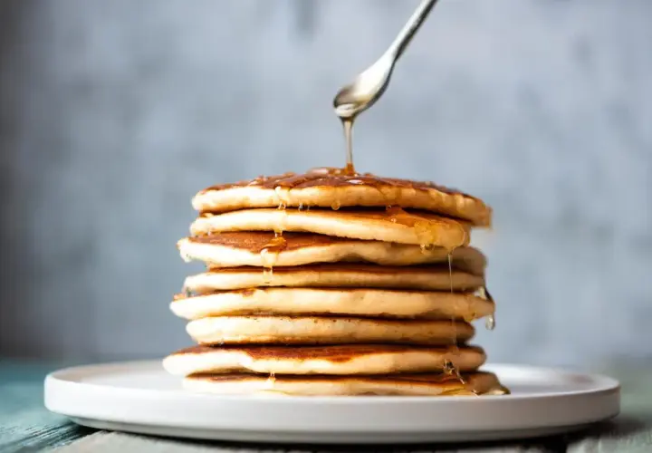 Una pila de tortitas sin mantequilla, doradas y esponjosas, con sirope cayendo de una cuchara.