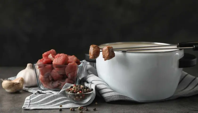 Preparación para un delicioso fondue de carne: trozos de carne cruda, ajo, granos de pimienta y una olla con caldo humeante.
