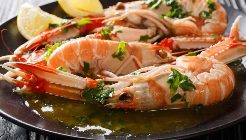 Cigalas a la plancha, jugosas y doradas, servidas con perejil picado y gajos de limón en un plato oscuro.