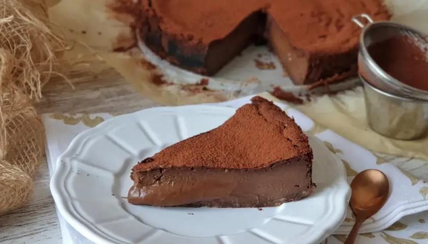 Una porción de tarta de chocolate, cubierta de cacao en polvo, reposa en un plato blanco. El resto de la tarta, también espolvoreada, se ve al fondo.