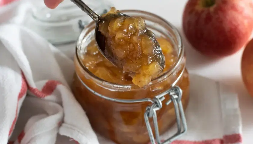 Cuchara sirviendo mermelada de manzana casera de un frasco. Manzanas rojas frescas al lado.
