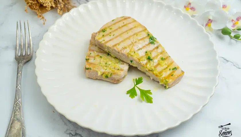 Dos filetes de atún a la plancha, con marcas de parrilla y aderezados con ajo y perejil, servidos en un plato blanco con un tenedor.