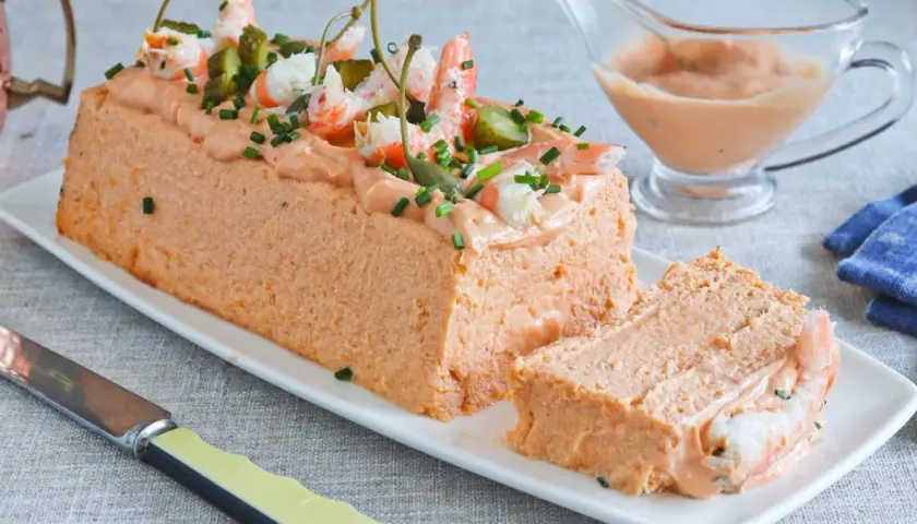 Pastel de pescado decorado con gambas, pepinillos y alcaparras. Servido en una bandeja blanca con una porción cortada.