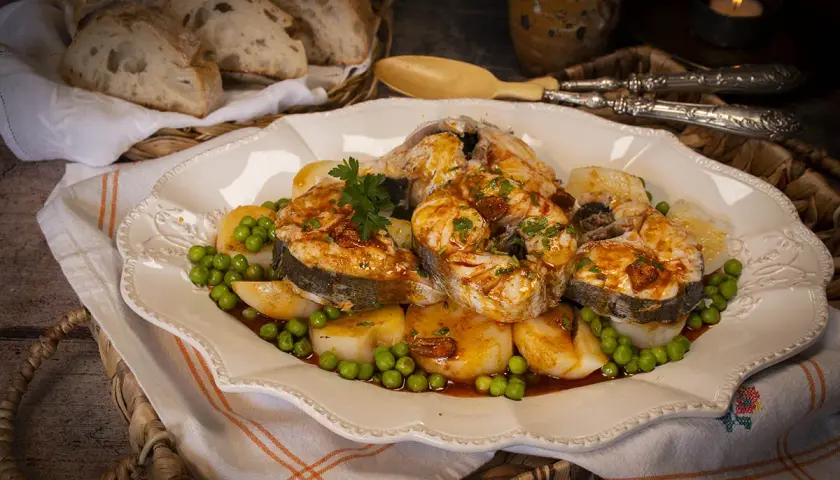 Merluza a la gallega arguiñano, jugosos trozos de pescado con patatas y guisantes, servidos en una fuente decorada.