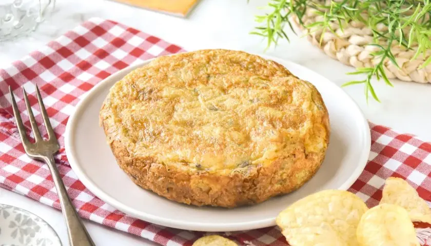 Tortilla con patatas de bolsa, dorada y jugosa, servida en plato blanco sobre mantel de cuadros rojos. Patatas fritas crujientes alrededor.