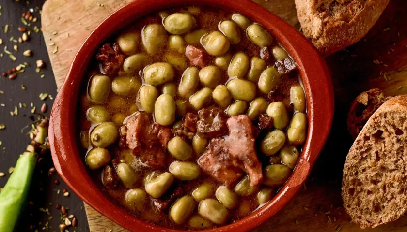 Un cuenco de barro con habas a la catalana, trozos de tocino y pan rústico al lado.