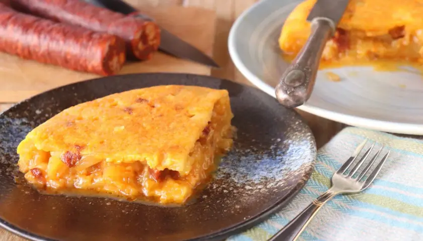 Deliciosa tortilla de chorizo, jugosa y dorada, servida en plato oscuro. Chorizos crudos al fondo.