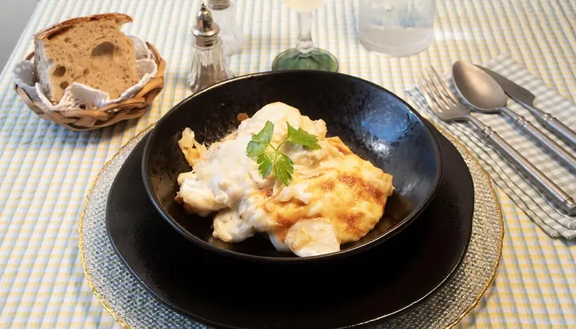 Delicioso bacalao a la nata gratinado, servido en un cuenco negro, acompañado de pan rústico y cubiertos.