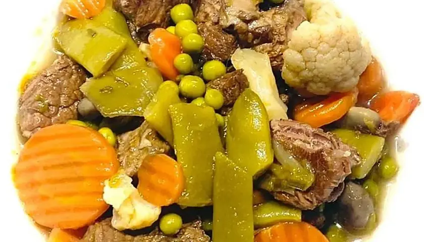 Ternera a la jardinera con trozos de carne tierna, zanahorias, guisantes, pimientos verdes y coliflor en un plato blanco.