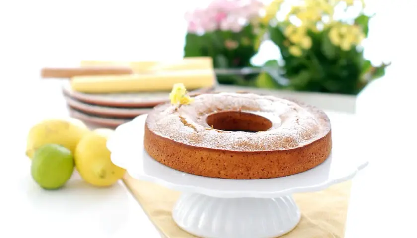 Bizcocho de limón Thermomix esponjoso y dorado, espolvoreado con azúcar glas. Limones y lima frescos al lado.
