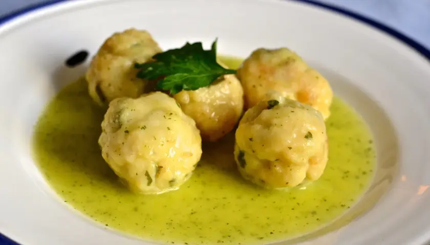 Cinco albóndigas de merluza caseras flotan en una salsa verde cremosa, adornadas con una ramita de perejil fresco.